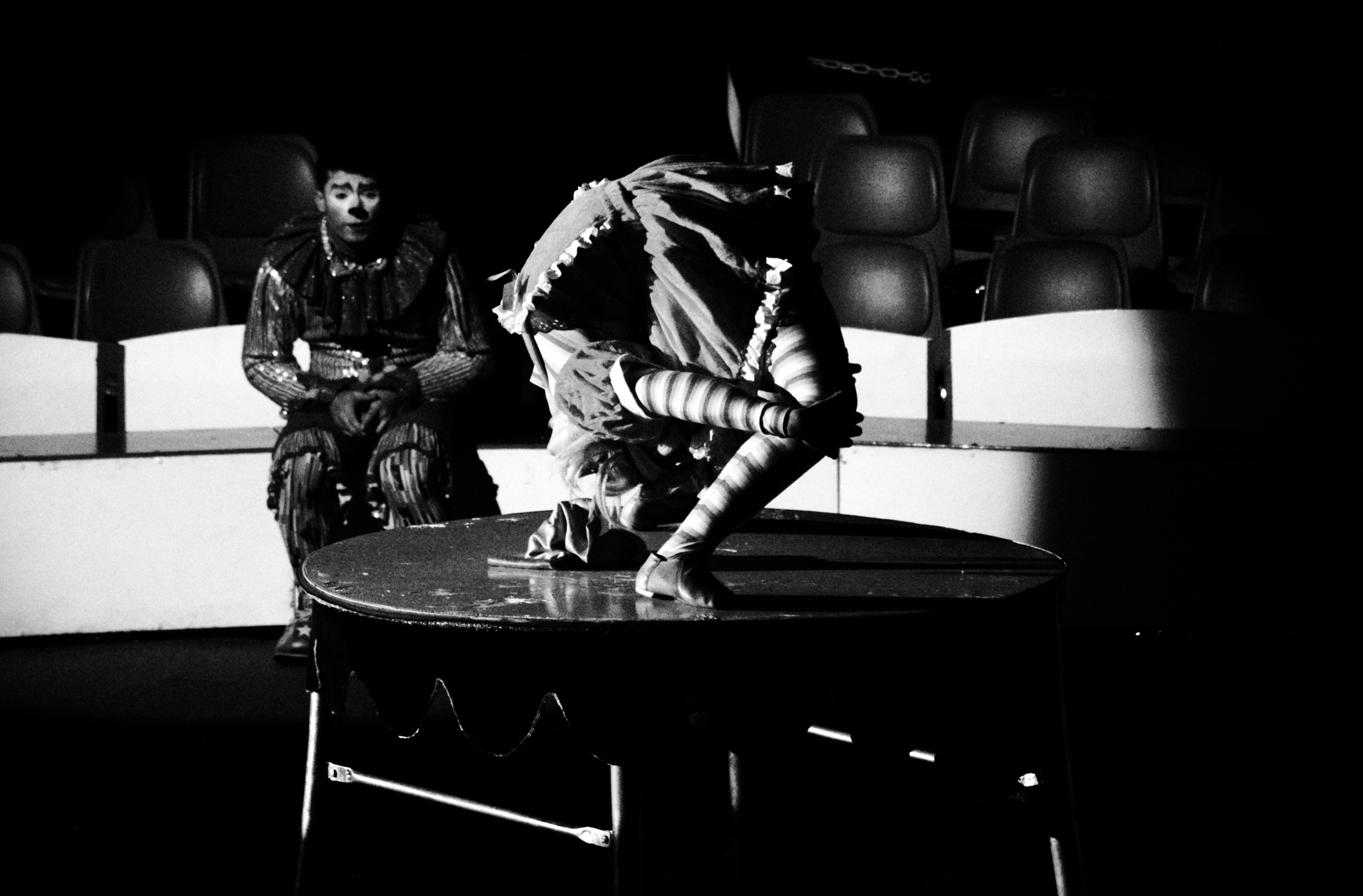 One performer watches another contort on a circus table, empty seats behind